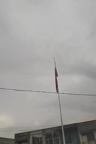 The media file shows a tall, white flagpole with a red flag with white lettering flying from it. The flag is positioned in the center of the frame, against a cloudy, overcast sky. Below the flagpole and the flag, a row of buildings is visible. These appear to be shop fronts, with awnings or covered walkways extending from them. Some of the buildings look slightly weathered. A black wire runs horizontally across the frame, just above the buildings and below the flag. The overall scene suggests an outdoor setting during the day, with the weather being cloudy. The location is identified as Keudegeudong, Indonesia. Visible text on the buildings includes "PUSEK" and " Hannochs" on signs, and other indistinct writing. The presence of the flag on a flagpole indicates a public space where a flag is flown, possibly for official purposes or as a business or organizational marker.