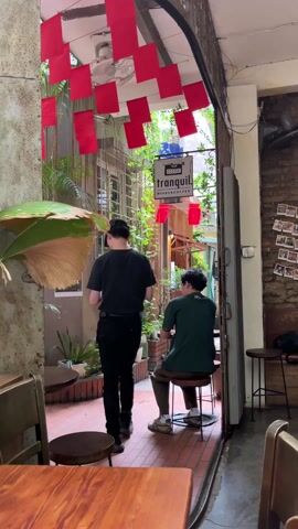 The interior of a café is captured, showing a doorway leading to an outdoor area, where a man sits on a stool, facing away from the camera. He is wearing a green shirt and appears to be smoking. A second man, dressed in black, walks through the doorway into the outdoor space. A sign above the entrance reads "tranquil. BOOKS & COFFEE". Red square decorations are hung above the doorway, and plants are placed around the outdoor area. Inside the café, wooden tables and chairs are visible, along with a brick wall adorned with photographs.