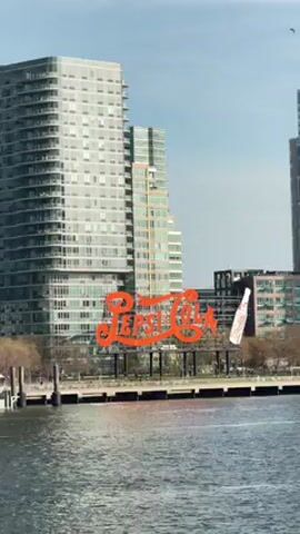 A bright daytime shot captures a wide urban waterfront landscape, centered around the iconic "PEPSI-COLA" sign. The large, historic sign is rendered in bold orange-red script, with a classic white Pepsi bottle silhouette positioned to its right. It stands prominently on a strip of land by the water's edge.

Behind the sign, the background is dominated by several tall, modern buildings, characterized by their numerous windows and balconies, suggesting residential or mixed-use high-rises. The architectural style appears contemporary. In the foreground, a body of water, likely a river or bay, stretches across the frame, showing slight ripples on its surface. To the left, a small pier or dock structure with pilings extends into the water. The land immediately behind the sign features sparse, bare trees, indicative of late winter or early spring.

The sky is a clear and bright light blue, suggesting fair weather conditions. The overall lighting is strong and even, illuminating the buildings and the sign clearly. No people or specific activities are visible, presenting a static, picturesque view of this well-known New York City landmark and its surrounding urban development.