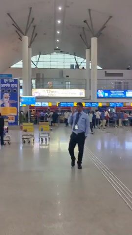 The video shows a wide shot of a busy airport terminal. The ceiling is high and white with exposed beams and numerous lights. The floor is polished and reflective. There are several rows of luggage carts visible. People are walking in various directions, some with luggage. To the right, a man in a blue uniform with a lanyard is walking and talking on his phone. Behind him, there are check-in counters with signs and people queuing. A large blue advertisement with text in Hindi is visible on the left wall. The overall impression is of a bustling travel environment.