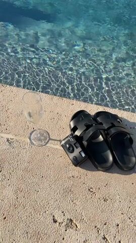 A high-angle shot shows a poolside scene with a champagne flute, a small black disposable camera, and a pair of black slides resting on the light-colored concrete edge of a swimming pool. The water in the pool is clear and blue, with sunlight creating ripples and reflections on its surface. The objects are positioned in a line from left to right: the flute, the camera, and then the slides.