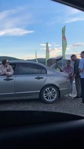 A group of three people are gathered near the open trunk of a silver sedan. One woman is reaching into the trunk, while a man stands next to her, and another man stands behind him. Two green flags with a red logo are visible in the background, along with a cloudy sky and distant hills. The video then shifts to focus on the back of a young child with curly dark hair, seen from behind, sitting inside a car. The child is wearing a light-colored shirt.