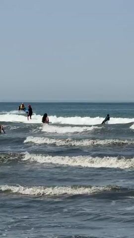 A beach scene with gentle waves rolling in. In the foreground, the dark water is choppy. In the middle ground, white-capped waves break closer to shore. A person with dark hair is visible in the water. Further out, a jet ski with two people, one in a yellow top, moves across the scene. Two individuals are on surfboards in the water, one paddling and the other standing. The sky is clear and pale blue.