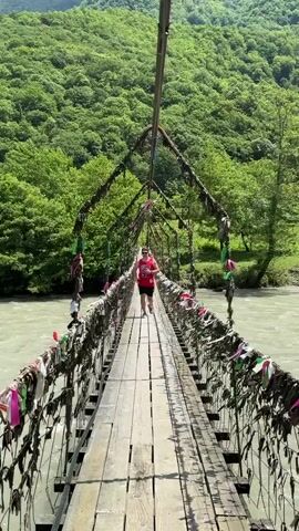 A man walks on a rustic wooden suspension bridge spanning a wide, light-colored river. The bridge's wire railings are adorned with numerous colorful ribbons and pieces of cloth, suggesting a tradition or a popular tourist site. The surrounding landscape features steep, lush green hills and mountains covered with dense trees under a bright, sunny sky. The man, wearing a red sleeveless jersey with "BULLS 91" printed in white on the front, black shorts, and sunglasses, walks confidently towards the camera. The river beneath the bridge flows swiftly, and the overall scene conveys a serene yet active outdoor setting, likely in a mountainous, forested region.