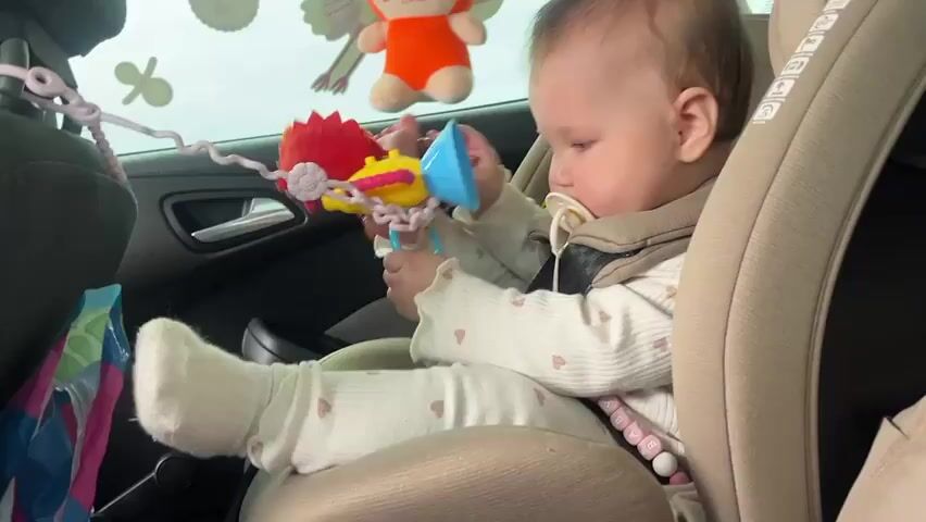 The video shows a baby sitting in a car seat. The baby is wearing a white, long-sleeved outfit with heart-shaped patterns and a pacifier in their mouth. They are secured in the car seat with a harness. The baby is playing with a colorful toy that hangs in front of them. The toy has various shapes and colors, including blue, yellow, and red. In the background, there is a car window with cartoon-like stickers and a plush toy attached.  A colorful bag is partially visible next to the car door. The car interior is clean and well-maintained.