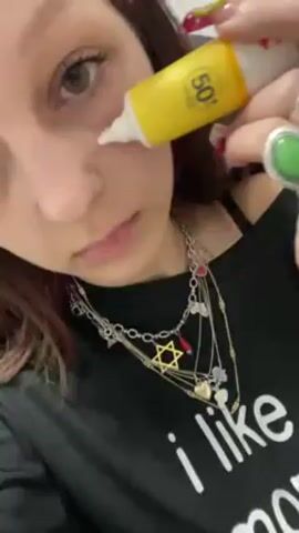 Here is a description of the video:
The video shows a young woman applying sunscreen to her face. She has light skin, light eyes, and brown hair with a pink streak. She is wearing a black t-shirt with white writing and several necklaces, including one with a Star of David pendant. She's also wearing a green gemstone ring. She uses a small, yellow tube of SPF 50 sunscreen and applies it in streaks on her cheek and forehead. Music plays in the background. The focus is on the woman's face as she applies the sunscreen.