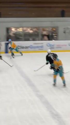 A hockey game is in progress on an ice rink. Several players in uniform are visible, some in black and green jerseys, others in yellow and teal jerseys. One player in a black and green jersey with the number 8 on their back is skating towards the right side of the frame. Another player in a yellow and teal jersey is skating towards the left. A referee in black and white stripes is standing in the background. Spectators are visible in the bleachers above the ice. The environment appears to be an indoor ice arena with wooden seating and glass barriers.