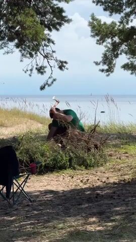 A person is crouched down near a pile of green branches on a sandy beach. They are holding an axe and appear to be chopping or manipulating the branches. In the background, there is water, a hazy sky, and trees with pine needles. In the foreground, there is a folding camp chair with a dark jacket draped over it, and a red cup. Further back, there is a tent. The scene is illuminated by natural daylight.