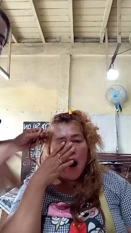 A woman with blonde, messy hair is sitting in front of the camera, her mouth open in a surprised expression. She is wearing a black and white checkered shirt with a colorful cartoon print on the chest. Her right hand is raised, touching her cheek near her eye. To her left, another person, whose face is not fully visible, is working on her hair, seemingly pinning a section of it. The background appears to be an indoor setting, possibly a salon, with a wooden ceiling, a fan mounted on the wall, and a sign with text that is partially obscured. The lighting suggests an overhead light source.