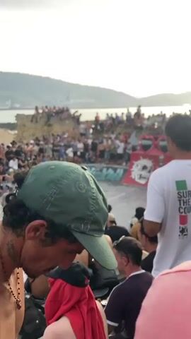 A large crowd watches a skateboarder perform a trick at a skate park. The skate park appears to be located near the coast with hills and a body of water in the background. Many people are sitting and standing around the skate park, some are taking pictures. There is a ramp with a large pair of red sunglasses at the top. The skateboarder is in mid-air performing a trick. The time of day appears to be sunset as the sky is hazy and orange.
