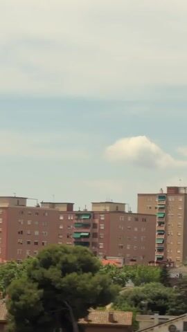 The video shows a view from a moving vehicle, likely a train or car, looking out at a city landscape. The view is a mix of buildings, trees, and sky. In the foreground, the vehicle's interior, including a window and some structural elements, are visible. The outside scene is blurred due to the motion. Several apartment buildings of varying heights and colors, with some featuring green awnings, are prominent. Trees and greenery are interspersed between the buildings and in the foreground. The sky is partly cloudy. There are streetlights visible on poles, and some construction scaffolding is also present. The overall impression is of a sunny day in an urban or suburban environment.