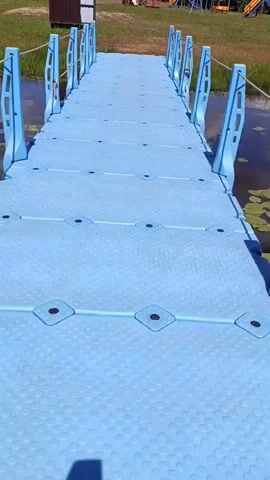 A first-person perspective video shows a bright blue modular floating dock extending over a body of dark water. The dock is made of interconnected hexagonal plastic modules, each with raised circular textures and diamond-shaped black inserts. Blue vertical supports with rope railings line both sides of the dock. The dock appears to be leading towards a grassy bank. In the background, on the bank, there is a brown shed, some trees, and a children's playground with slides and swings under a blue sky with scattered white clouds. The camera moves forward, giving a clear view of the dock's surface and the surrounding environment.