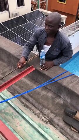 A bald Black man wearing a grey jumpsuit over a white undershirt stands on a rooftop next to a swimming pool. He is holding a bottle of water and appears to be working. A blue rope is stretched across the rooftop in front of him, and there are solar panels visible in the background. The man is looking down at the rooftop.