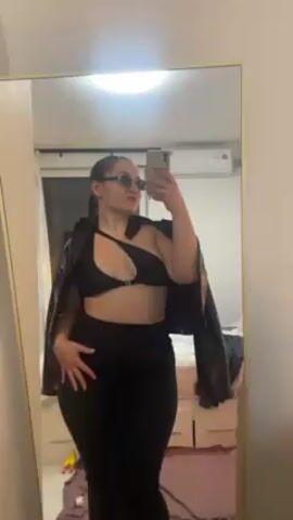 A woman is taking a selfie in a mirror. She is wearing a black asymmetrical top, black pants, and a brown leather jacket draped over her shoulders. She also has round sunglasses on. The background shows a room with a bed and an air conditioner. The bed is covered with a pink sheet and some clothes are scattered on it. The lighting is bright. Music is playing in the background.