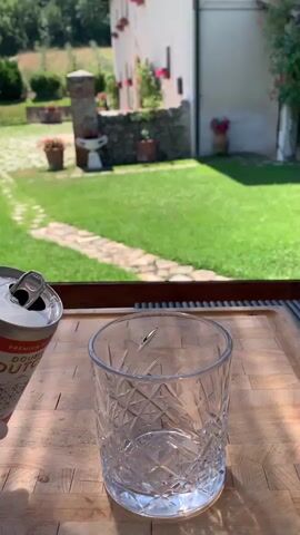 In this video, a hand holds a can of "DUTCH" premium ginger ale and pours it into a clear, faceted glass filled with ice. The drink fizzes as it fills the glass. The scene appears to be outdoors, with a well-maintained green lawn and a rustic stone building with potted plants in the background. The lighting suggests it is daytime, possibly late morning or early afternoon. The setting hints at a warm, pleasant atmosphere. The building's architecture and the surrounding landscape are characteristic of rural Italy, specifically the Tuscany region, with the location context provided as Castiglione d'Orcia. The focus is on the simple act of preparing a refreshing beverage in a picturesque environment.