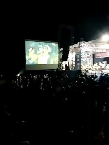 The video captures an outdoor evening concert from the perspective of a person within a large crowd. In the foreground, the crowd is densely packed, with many individuals silhouetted against the stage lighting. Their faces are mostly obscured by darkness, but some are holding up phones, suggesting they are recording or taking pictures. Further back, a stage is visible, adorned with lights and banners, where a band or musical group is performing. The stage is illuminated by bright spotlights. To the left of the stage, a large projection screen displays an image, possibly of the performers or related content. The overall environment is dark, indicating it is nighttime, with the stage and screen providing the primary sources of light. The atmosphere appears to be lively and engaged, with the audience gathered to watch the performance.