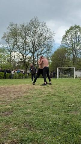 Two shirtless men in dark pants are wrestling on a grassy field. They are closely grappling with each other. A small crowd of people stands around the perimeter of the field, watching the wrestling match. There is a soccer goal visible on the right side of the frame, and trees surround the field. The sky is overcast.