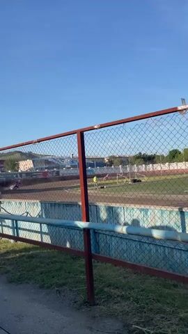 The video shows a motorcycle race on a dirt track. Several motorcycles with riders are visible on the track. In the background, there are spectators in a stadium, buildings, trees, and a clear blue sky. The camera is positioned behind a fence, which partially obstructs the view. There are also two boys walking in the foreground on a path next to the fence.