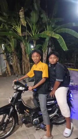 The video shows two young males outdoors at night, with tropical foliage in the background. The first male, wearing a yellow and black shirt and gray pants, is seated on a black motorcycle. He is looking at the camera and smiling. The second male, wearing a black shirt and white pants, stands next to the motorcycle, resting his arm on the seat. He is also looking towards the camera. The first male then turns and rides the motorcycle away. The video captures the scene from various angles as the motorcycle moves.