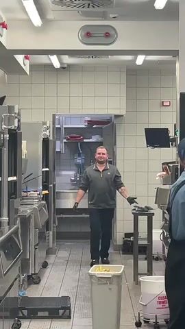 A man in a dark grey uniform with a gold logo on the left side walks towards the camera in a commercial kitchen. He is wearing black gloves and dark pants. The kitchen has tiled walls and floors and stainless steel equipment. There is a trash can with a white bag liner in the foreground. The man smiles and waves at the camera.