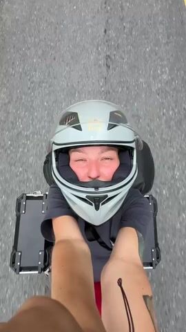 The video captures a first-person perspective of a motorcycle ride through a mountainous landscape in Minh Sơn Commune, Vietnam.

The initial shot at 0:00 shows the person filming, wearing a light grey full-face motorcycle helmet with "Royal" visible on the front. Their arms are extended, holding the camera, and one arm displays a tattoo. Black motorcycle side cases are visible behind them on what appears to be a motorcycle seat. The ground is asphalt.

At 0:01, the view shifts to the side, revealing a winding asphalt road bordered by red and white striped concrete barriers. Lush, verdant mountains, thickly covered in trees, dominate the background under a cloudy sky.

From 0:02, the camera is positioned behind a different motorcycle helmet, also light grey, featuring a neon green accent on top and a black and white graphic design with partial text "THE" and a stylized "H" visible. The rider's hand is seen on the handlebar, navigating the winding mountain road. The majestic green mountains and valleys stretch into the distance, partially obscured by clouds. Red and white striped barriers continue to mark the edge of the road.

The final frame at 0:04 provides a broader view of the scenic mountain road. Two red and white striped barriers are in the foreground, framing the dramatic landscape of towering green mountains under a cloudy sky, suggesting a high-altitude ride.