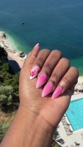 The video shows a close-up of a left hand with stiletto-shaped, bright pink acrylic nails. The thumb and index finger are visible, followed by the middle finger, ring finger, and pinky finger which are spread out. The nails on the thumb, middle, ring, and pinky fingers are a solid bright pink color with a thin silver line at the cuticle. The index finger nail has a gradient pink and white design with a small pink floral accent near the tip. The hand is positioned against a backdrop of a turquoise body of water, a beach with lounge chairs, a swimming pool, and lush green vegetation, suggesting an outdoor, possibly tropical, location. The lighting is bright and sunny.