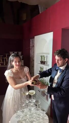 A bride and groom are pouring champagne into a tower of champagne glasses. The bride, on the left, is wearing a white dress, and the groom, on the right, is wearing a dark blue tuxedo with a blue bow tie. The tower of glasses is arranged on a round table covered with a white tablecloth. The room has red walls and a white shelving unit behind the couple.