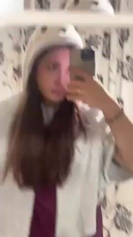 A woman films herself in a mirror using a cell phone. She is wearing a white, hooded jacket with a cartoon owl face on the hood. She is wearing a dark pink dress or shirt. She is wearing a gold ring on her left hand and a bracelet on her right hand. Her hair is long and brown. The background is a bathroom with patterned wallpaper.