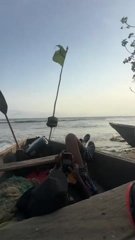 The image captures a peaceful coastal scene in Limbé III, Cameroon, likely during early morning or late afternoon due to the soft, warm light. A person is lying relaxed inside a wooden fishing boat, which is beached on the sand. The individual, wearing patterned shorts and flip-flops, is focused on a smartphone held in their hands.

The boat is equipped with an outboard motor at the stern, a paddle leaning against the side, and fishing nets or ropes stored within. A tall pole topped with a small, light green flag stands prominently from the boat's interior. In the background, the ocean extends to the horizon, with gentle waves breaking on the shore under a clear, light blue sky that fades to a soft yellow-white near the horizon. Part of a second wooden boat is visible on the right, further down the beach, and some sparse tree foliage borders the scene on the far right. The setting suggests a tranquil moment of leisure by the sea.