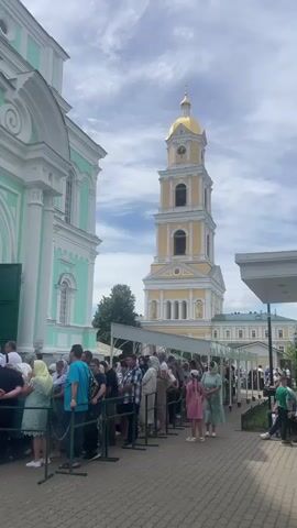 The video shows a long line of people standing outside a building. The building is green and white with a tall yellow tower with a gold dome. The people are all facing the building and appear to be waiting to enter. The ground is paved and there are trees in the background. The sky is cloudy.
