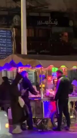 Here's a description of the video:

The video shows a street scene at night. A brightly lit food stand is the focal point, with several people gathered around it. The stand has blue, green, and other colored lights illuminating its canopy and sides. People are standing and appear to be ordering or receiving food. There is a sign written in Arabic visible above the stand.

The camera pans out to show the street. Cars are driving by, and pedestrians are walking along the sidewalk. There are trees and buildings in the background, illuminated by streetlights. The overall impression is of a typical nighttime street scene in a city, centered on the activity around the food stand.