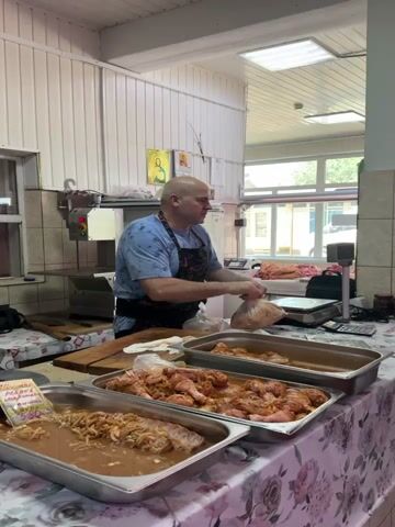 A bald man wearing a blue t-shirt and a dark apron with chili pepper designs is standing behind a butcher's counter. He is holding a plastic bag filled with meat and appears to be tying it. In front of him, on the counter, are three large metal trays filled with raw chicken pieces in a marinade. To his right, there is a scale and a calculator. Behind him, there is a commercial meat grinder or saw, a refrigerator, and religious icons on the wall. The environment appears to be a butcher shop or a food preparation area.
