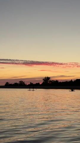 A drone shot captures a serene sunset over a body of water. Silhouettes of trees line the horizon, framing the pastel hues of the sky. Several people are visible on the water, engaged in stand-up paddleboarding and kayaking. The calm surface of the water reflects the warm tones of the sunset, creating a peaceful and tranquil atmosphere.