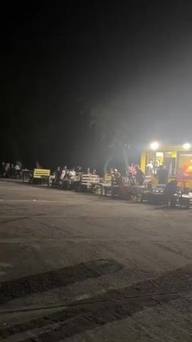 The video shows a group of people sitting at outdoor tables at night. There is a yellow building in the background with lights on. The people appear to be eating or drinking. The ground is dark and appears to be dirt or gravel. The lighting is dim and hazy.