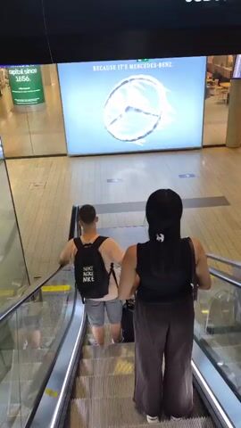 A man and a woman are riding an escalator down. The man, on the left, has a black backpack with "NIKE" written on it multiple times and is pulling a black suitcase. He is wearing a tan t-shirt and grey shorts. The woman, on the right, is wearing a black tank top and brown pants and is also pulling a black suitcase. They are both facing away from the camera. In the background, a large illuminated advertisement displays the Mercedes-Benz logo and text that reads "TESTED AT 40°C. BECAUSE IT'S MERCEDES-BENZ." The environment appears to be an airport or a large public building with tiled flooring. There are directional signs in the background.