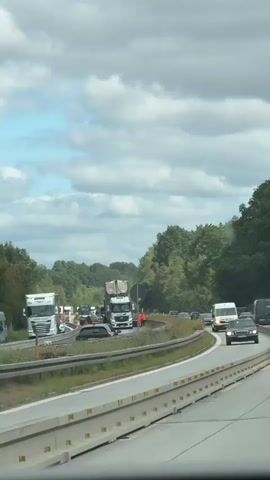 The video shows a highway with heavy traffic congestion and what appears to be an accident scene. Several large trucks, including a white semi-truck with a trailer and a construction vehicle, are stopped on the side of the road. There are also cars and vans present. Some individuals in orange vests, likely emergency personnel or road workers, are visible near the trucks, suggesting some form of intervention or cleanup is underway. The highway is bordered by trees and vegetation on one side, and a barrier separates the lanes of traffic. The sky is cloudy. The overall scene indicates a disruption to normal traffic flow due to an incident.