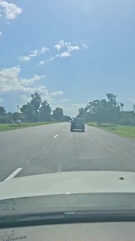 The video shows a view from the inside of a car driving on a two-lane road on a sunny day. The road is paved and has white lane markings. There are trees and vegetation on both sides of the road, and a few scattered clouds in the blue sky. Several cars are visible on the road ahead. One car is in front of the camera car, and another car is approaching from the opposite direction. The sun is shining brightly, casting shadows on the road. The overall impression is that of a typical drive through a rural or suburban area on a clear day.