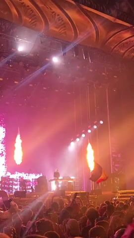A DJ is performing on stage at a concert. The stage is lit with bright pink and purple lights, and there are large screens displaying visuals on either side. A Belgian flag is being waved by someone in the audience. The crowd is energetic and appears to be enjoying the performance. There are also flames erupting from the stage.