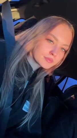 A young woman is in a vehicle, possibly a bus or car, wearing a black jacket with the word “FASHION” on it. She is fair-skinned, with blonde hair and blue eyes. She looks directly at the camera and slightly puckers her lips. There is natural sunlight hitting her face. The vehicle is moving and the background is dark.