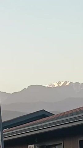 A video shows a distant mountain range with snow-capped peaks under a clear sky. In the foreground, parts of buildings and streetlights are visible. The camera pans slightly to the left.