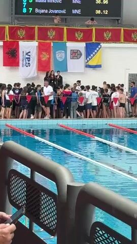 The video shows a crowd of young people gathered at the edge of a swimming pool. They appear to be at a swimming competition, as suggested by the lane markers in the pool and the scoreboard displaying names and times above. Several flags are displayed along the wall behind the crowd, including the flags of Montenegro and Bosnia and Herzegovina, as well as flags of other organizations. Some people are talking and gesturing, and some are holding or using their phones. The perspective is from behind a gray metal railing, likely a spectator area.