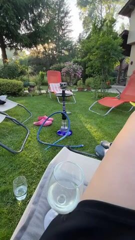 The video begins by showing a hookah set up on a grassy lawn in a backyard. There are several lawn chairs, a pair of pink flip-flops, and two glasses of white liquid on the lawn. The camera then pans to the right, revealing a house and more of the backyard, which is filled with trees and plants. The sun is setting, casting a warm glow over the scene. The video appears to be filmed from the perspective of someone sitting in a lawn chair.