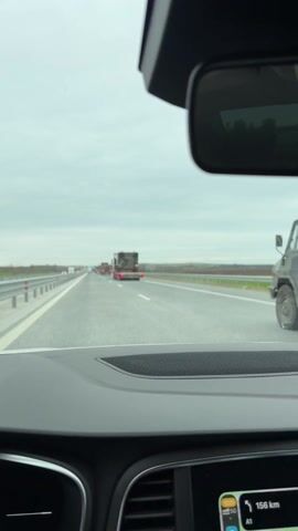 A car is traveling on a multi-lane highway with other cars in view and the camera is pointed at a convoy of several flatbed trucks carrying military vehicles. The sky is cloudy and the trees lining the sides of the highway appear leafless. The military vehicles being transported are covered in camouflage paint. The flatbed trucks transporting them are red.