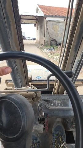 The video shows a first-person perspective of someone operating a forklift. The operator's hands are briefly visible on the steering wheel, and their feet are on the pedals. The forklift is moving forward on a dirt road between buildings. The forklift's frame and chains are visible, along with a dashboard with various controls. The surrounding environment includes buildings, debris, and scattered vegetation. The overall impression is of a working environment on a construction site.