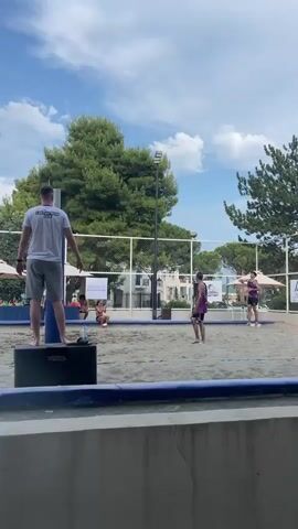 The video opens with a rear view of a man standing on a black speaker, looking towards a beach volleyball court. The court is made of sand, with a net dividing it in the middle. Two tall cypress trees are visible on the left, and a line of green trees is on the right. In the background, there are mountains and a cloudy blue sky. Two men are playing volleyball. One man in a white t-shirt and grey shorts serves the ball. The other man in a red and white tank top and dark shorts attempts to receive the serve. The server hits the ball high in the air. The receiver jumps and swings at the ball. The server is watching the ball. The first man is still standing on the speaker. The second man is now on the other side of the net, walking away from the camera.
