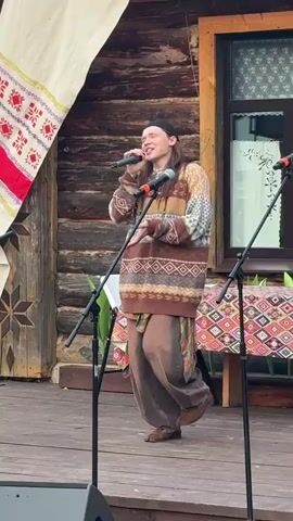 The video shows a person singing into a microphone on a wooden stage. The person is wearing a black beanie, a patterned sweater with brown, beige, and white colors, and baggy brown pants. A plaid shirt is tied around their waist. They are holding two microphones. The stage is set up outdoors, with a rustic wooden building in the background. A large banner with red and white embroidered patterns hangs to the left of the stage. A table with a colorful patterned tablecloth is visible behind the singer. Two microphones on stands are positioned in front of the singer, and a portion of a speaker is visible in the foreground. The scene appears to be a performance or event taking place during the daytime. The singer's expression suggests they are engaged and enjoying their performance. The overall setting and decor, particularly the embroidered banner and the wooden building, hint at a cultural or traditional event in a rural location. The location context indicates this is taking place in Starye Bygi, Russia.