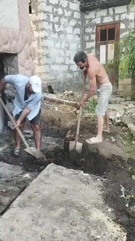 Two men are digging a hole in the ground with shovels. The man on the left is wearing a light blue shirt, dark shorts, a white cap, and sandals. The man on the right is shirtless, wearing light-colored shorts and sandals. They are in a yard with a building made of concrete blocks in the background. The building has a wooden door and window. There are some plants growing near the building. The ground is covered in dirt and some concrete slabs. The scene appears to be outdoors during the day. The weather seems to be warm as the man on the right is shirtless. The activity is manual labor, specifically excavation. There is no visible text in the media file.