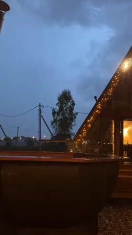 The video shows a heavy rain falling on a wooden, A-frame building decorated with string lights. The sky is dark and cloudy. There is a large, dark-colored vessel, possibly a hot tub, in the foreground. Other lights and buildings are visible in the distance. The camera pans upward to show the sky and the lights hanging under a wooden structure.