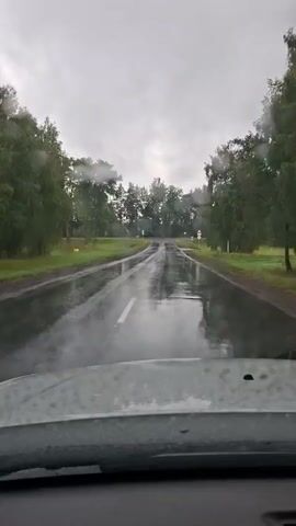 The video shows a view from inside a vehicle driving on a wet road. Raindrops are visible on the windshield, and the windshield wipers are activated at 00:01. The road is wet and reflective, with puddles and standing water in some areas. Trees line both sides of the road, and the sky is overcast and gray. There are a few road signs visible, including a speed limit sign indicating 20. The overall impression is of driving in rainy weather on a rural or suburban road.