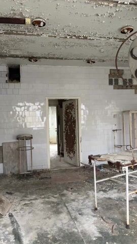 The video shows a decaying, abandoned operating room. The room has white tiled walls, but the ceiling's paint is peeling, and the floor is stained and littered with debris. A rusted surgical table is centrally located, along with a surgical light fixture hanging above it. A partially collapsed brick wall is visible on the right, and an open doorway leads to another room in the back. The room appears abandoned and damaged due to neglect.